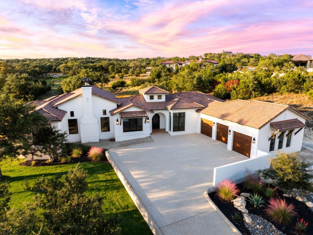 Aerial view of a Spanish Colonial custom home in Boerne, Texas, featuring white stucco, terracotta tile roofing, an arched entryway, and Hill Country landscaping at sunset.
