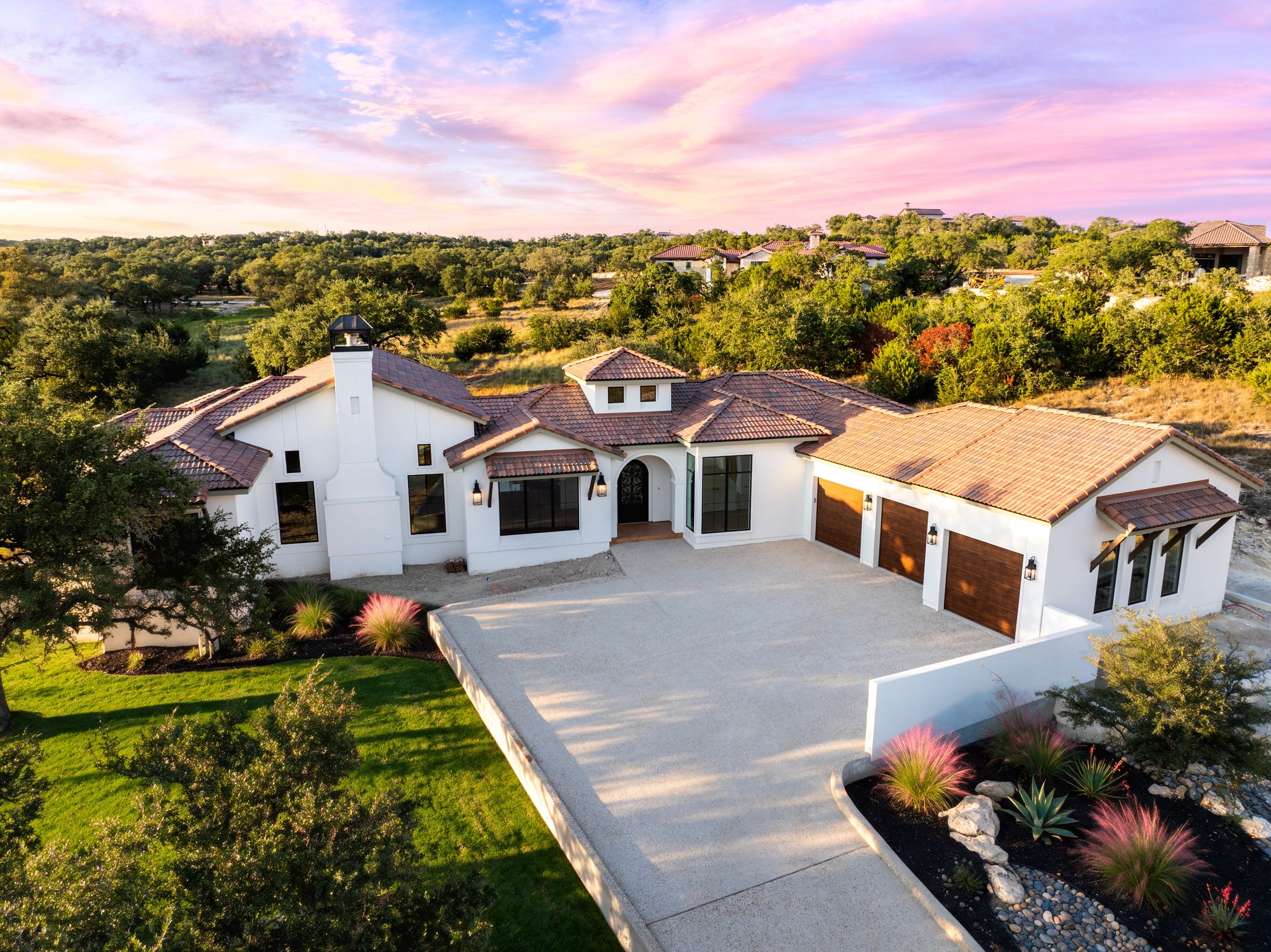Aerial view of a Spanish Colonial custom home in Boerne, Texas, featuring white stucco, terracotta tile roofing, an arched entryway, and Hill Country landscaping at sunset.